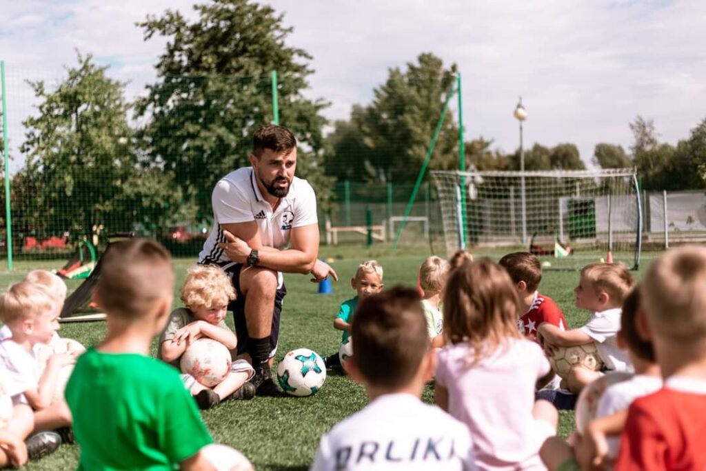 Trener tłumaczy siedzącym na murawie dzieciom zasady gry w piłkę nożną.