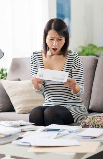 Kobieta z przerażoną miną czyta rachunek za prąd, siedząc na kanapie w swoim mieszkaniu. 