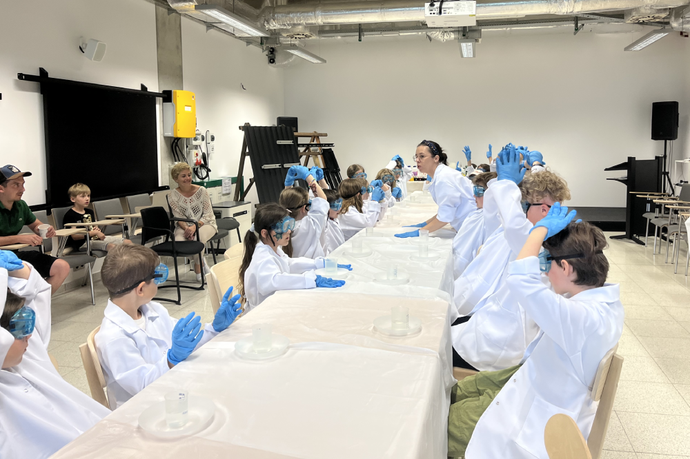 Grupa dzieci siedzi przy stole. Są w trakcie prowadzenia chemicznych eskperymentów. Nad nimi stoi pani, która tłumaczy zasady